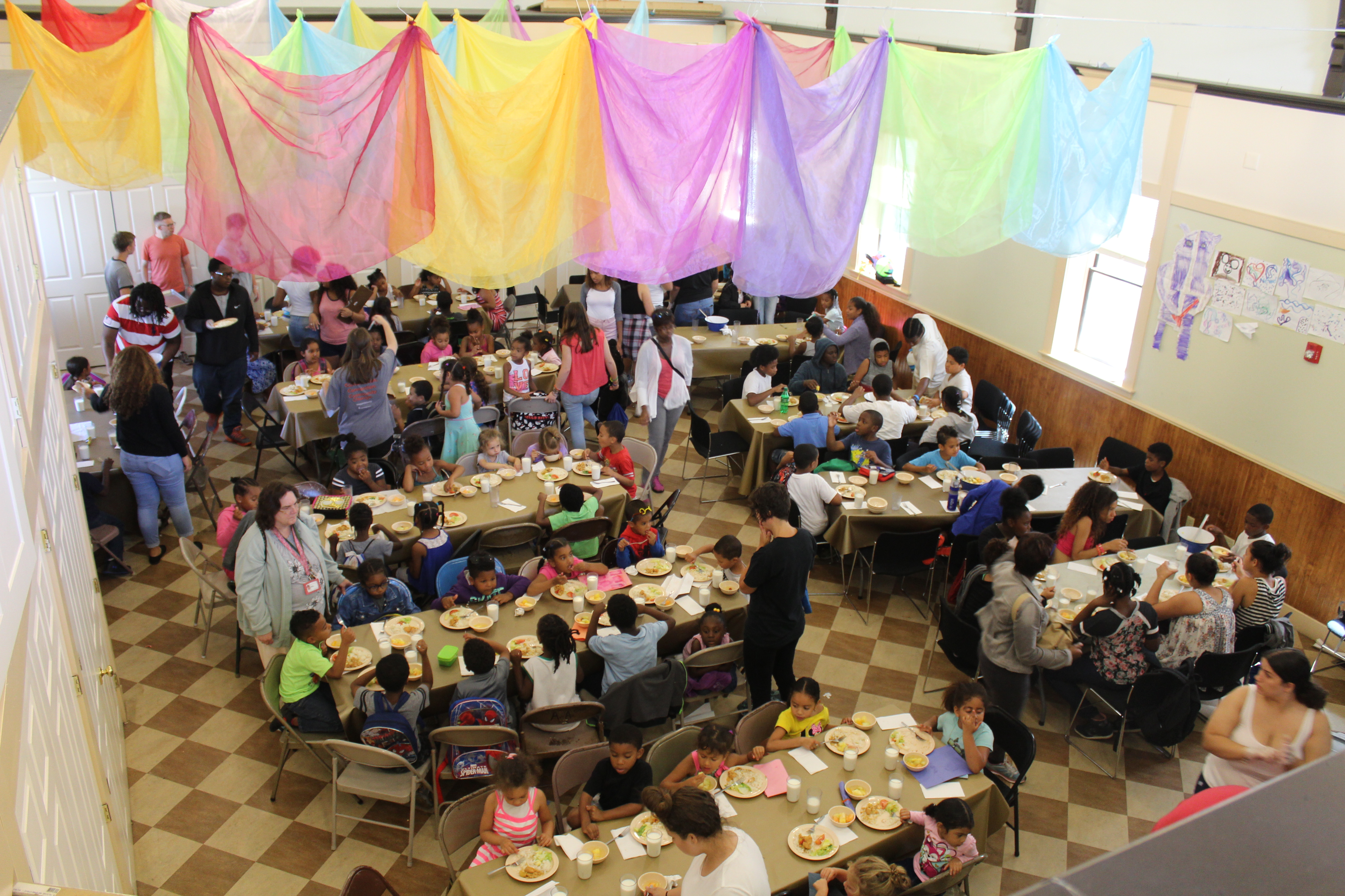 About one hundred kids sit at tables eating together, underneath beautiful rainbow fabric that hands from the ceiling above them.