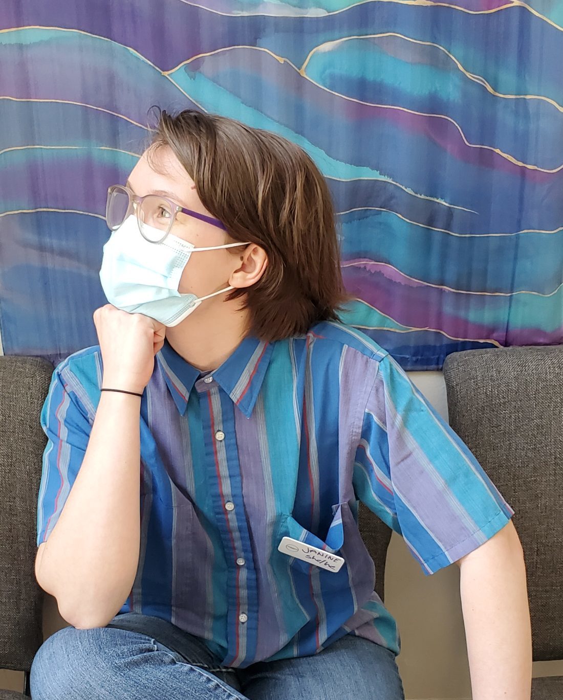 Janine, a white person with brown hair past their ears, purple glasses, and a surgical mask, sits with their chin perched on their fist. They wear a button-up shirt with vertical blue, purple, and red stripes.