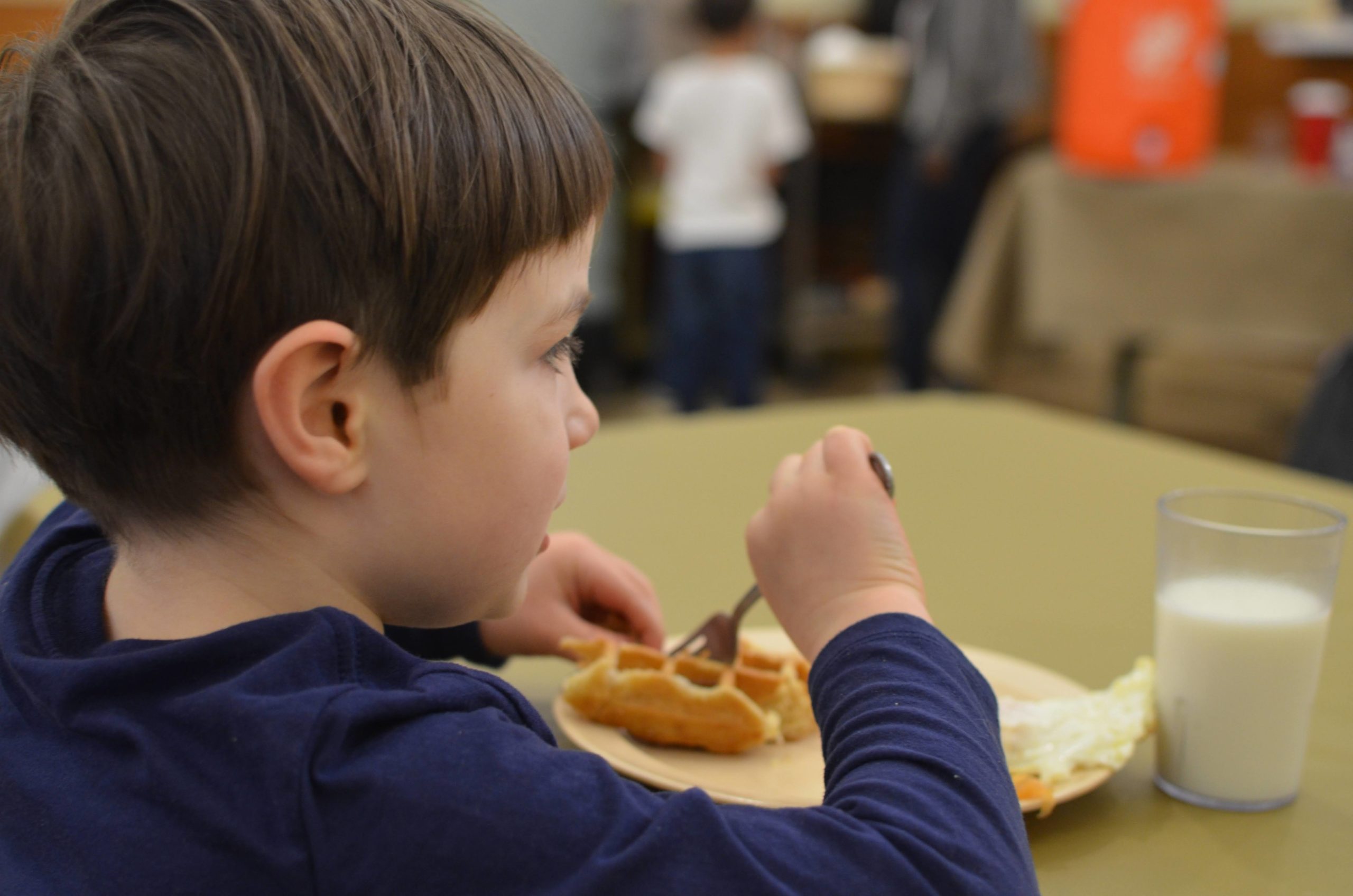 A 5-year-old kid sits at a table eating a waffle, eggs, and a glass of milk
