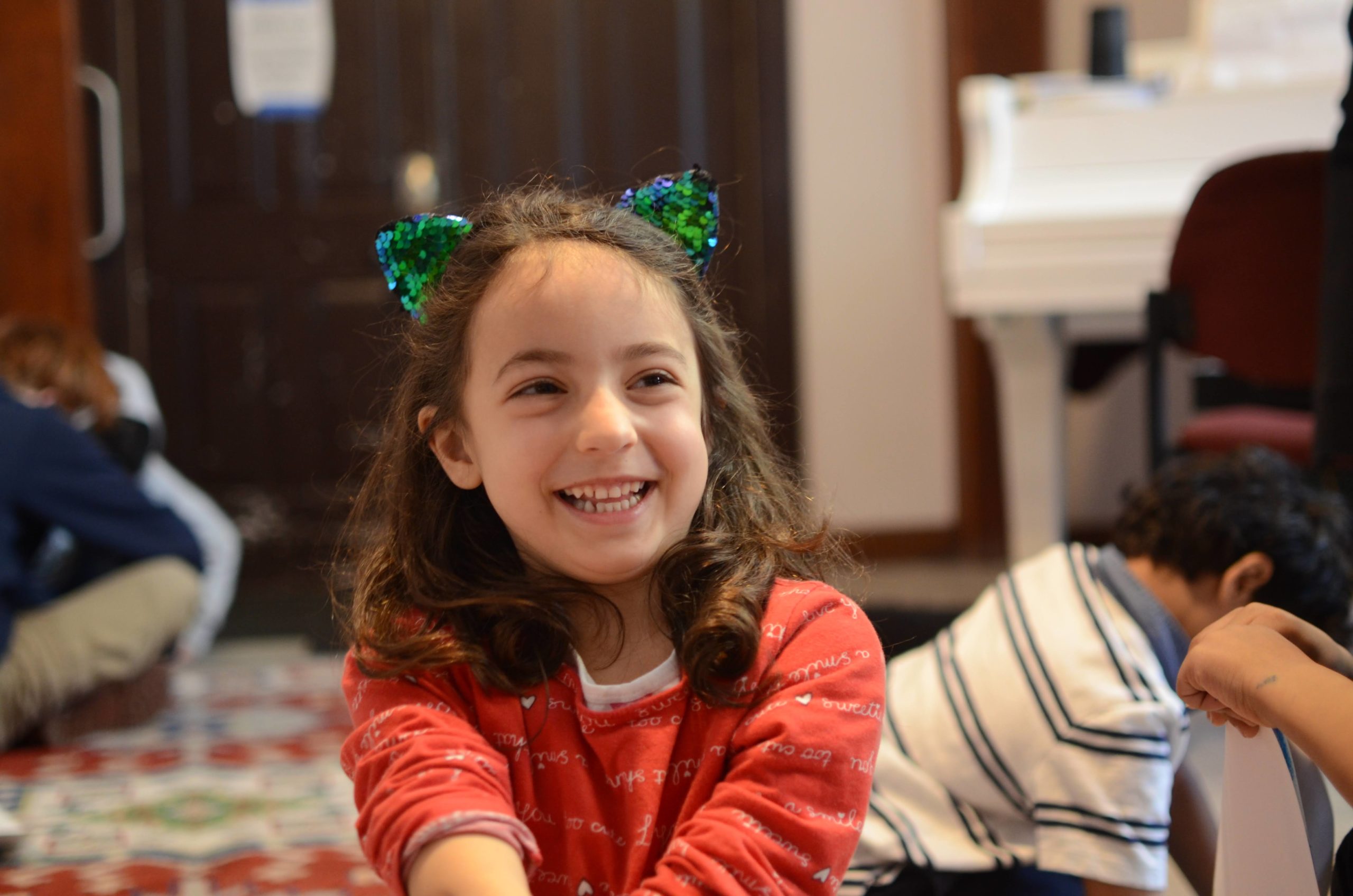 A 6-year-old kid wearing a sparkly cat-ear headband grins at someone sitting to the side of the camera
