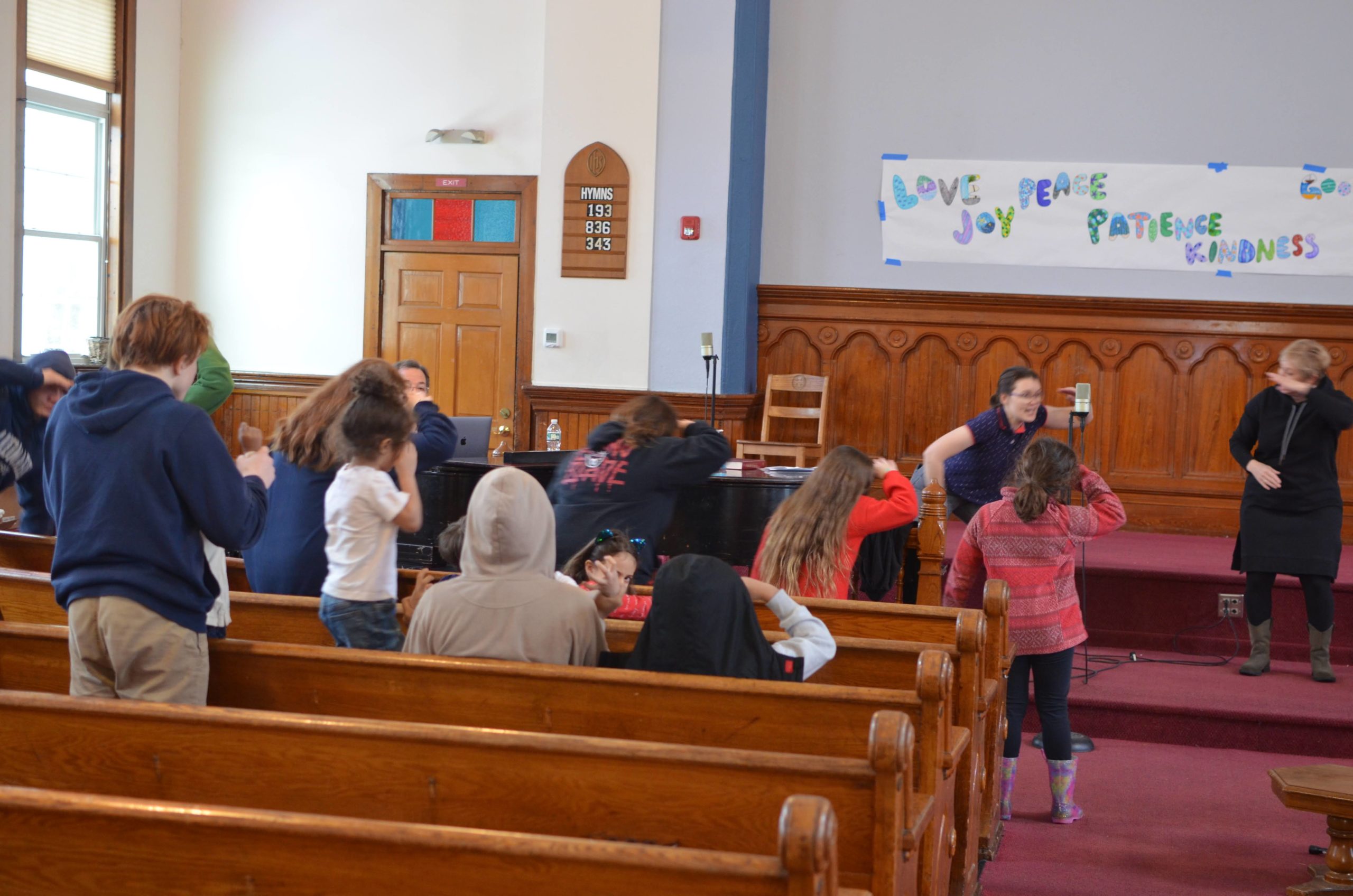 Camp director Janine and volunteer Alison lead kids in a song and dance in the sanctuary as they all place their hand above their eyes as if looking for something