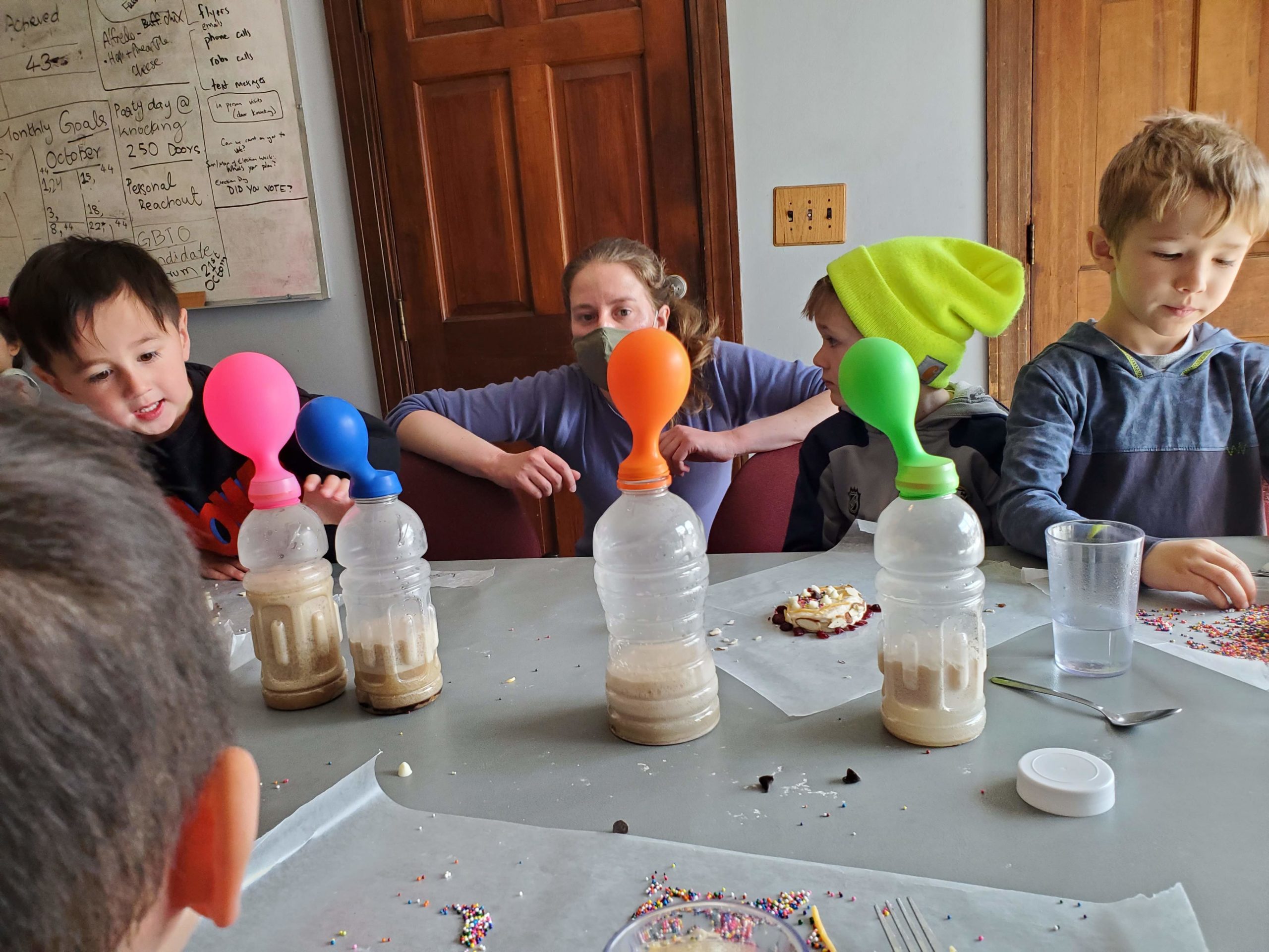 Science teacher Alexa crouches beside three young kids as they observe the gasses released by yeast and sugar in four gatorade bottles with balloons stretched over their openings.
