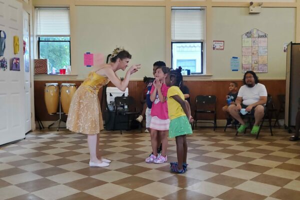 A ballerina in a dress and crown speaks with a small group of excited children