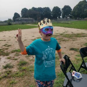 A child in a Summer Meals shirt, a paper crown, and a masquerade mask makes a peace sign at the camera