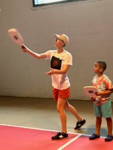 A grown-up demonstrates to a child how to serve in pickle ball on a pickle ball court