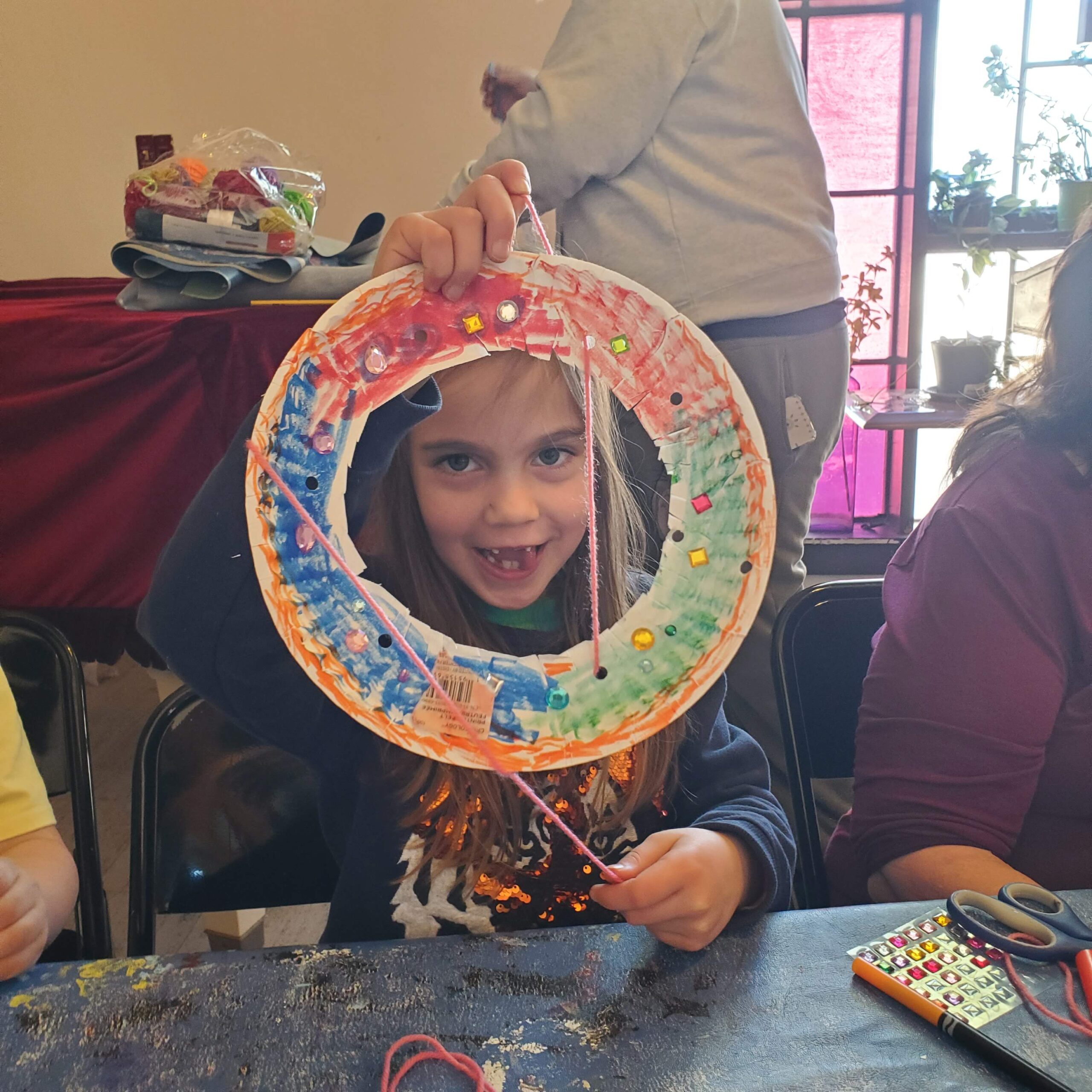 A young child holds up a colorful frame make out of a paper plate and smiles through it