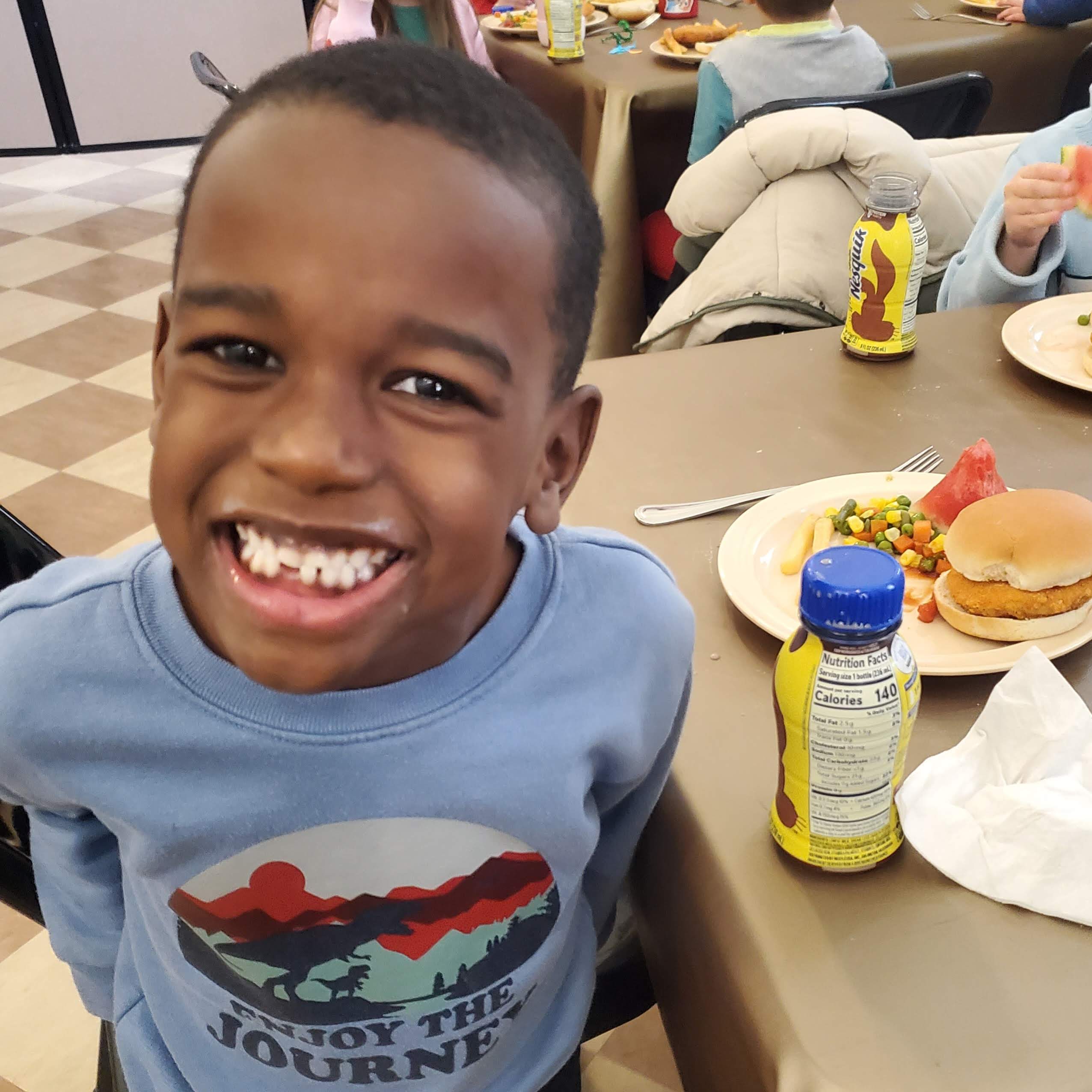 A young boy smiles beside a plate with a chicken sandwich, mixed veggies, watermelon, and chocolate milk.