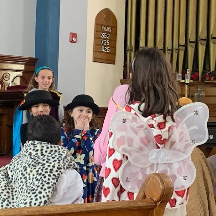 Children wearing bowler caps cover their mouths in surprise as children wearing angel wings approach